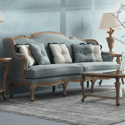 A classic living room showcases a Rococo Style Sofa adorned with exquisite wooden details and sumptuous cushions. This vintage blue centerpiece aligns beautifully with coordinating wooden lamps and a coffee table, all arranged on a neutral-toned rug against a light blue wall.