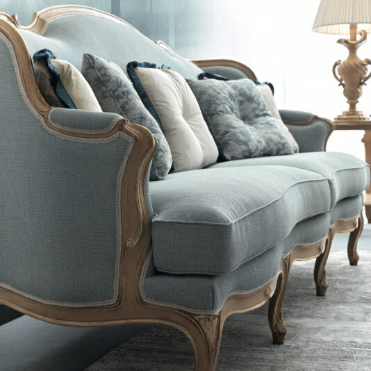 Rococo Style Sofa A stylish Rococo Style Sofa featuring blue upholstery and a wooden frame, accessorized with a mix of patterned and solid throw pillows. It sits atop a decorative rug next to an ornate lamp placed on a side table, creating a softly illuminated atmosphere.