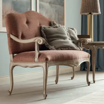 Small Rococo Style Day Sofa The Small Rococo Style Day Sofa, with its wooden frame and coral upholstery, is a charming vintage-style loveseat that sits gracefully in the living room. It comes with tufted details and is enhanced by two gray, fringed throw pillows. Nearby, a wooden side table and decorative lamp add to the elegant setting.