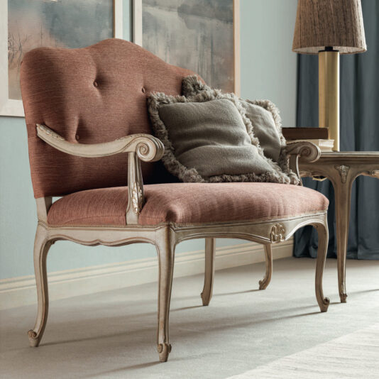 The Small Rococo Style Day Sofa, with its wooden frame and coral upholstery, is a charming vintage-style loveseat that sits gracefully in the living room. It comes with tufted details and is enhanced by two gray, fringed throw pillows. Nearby, a wooden side table and decorative lamp add to the elegant setting.
