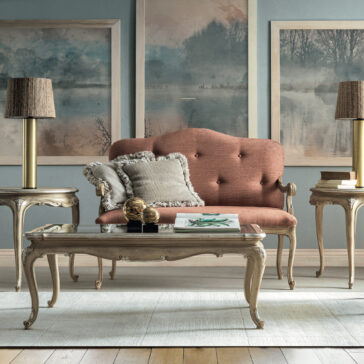 Small Rococo Style Day Sofa A cozy vintage living room includes a small Rococo style day sofa, adorned with textured cushions. It's flanked by elegant side tables with lamps. A glass-top coffee table sits on a light rug beneath three serene landscape paintings on the wall.