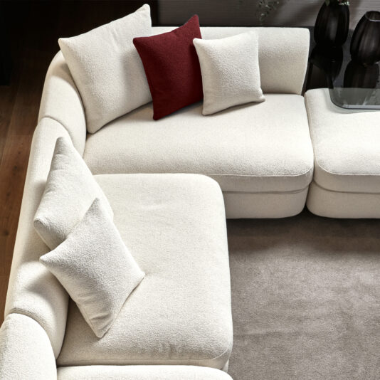 Large Modular Contemporary Corner Sofa In a living area, the Large Modular Contemporary Corner Sofa, a modern cream-colored sectional, is tastefully arranged. It features three cushions: two in cream that match the sofa itself and one in a contrasting deep red. The sofa rests on a light brown carpet, with two dark vases providing background decor.