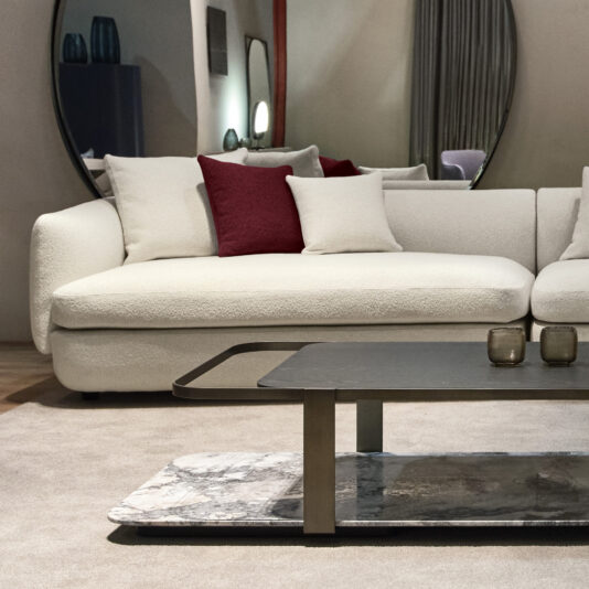 Large Modular Contemporary Corner Sofa A modern living room showcases the Large Modular Contemporary Corner Sofa, decorated with red and white cushions. In front of it stands a rectangular coffee table featuring a marble shelf, accompanied by two decorative vases. The cozy atmosphere is completed by a large round mirror and subdued lighting.