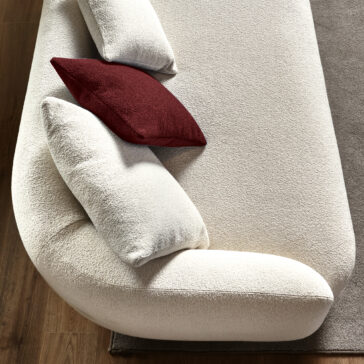 Large Modular Contemporary Corner Sofa From a top-down perspective, the Large Modular Contemporary Corner Sofa showcases a modern beige design with rounded edges. It comes with three plush cushions—two in beige and one in dark red—that sit gracefully on a wooden floor, with a gray rug partially visible underneath.