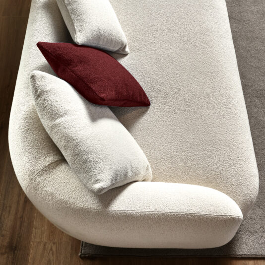 Large Modular Contemporary Corner Sofa From a top-down perspective, the Large Modular Contemporary Corner Sofa showcases a modern beige design with rounded edges. It comes with three plush cushions—two in beige and one in dark red—that sit gracefully on a wooden floor, with a gray rug partially visible underneath.