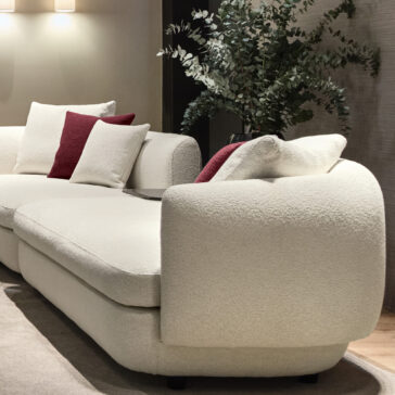Large Modular Contemporary Corner Sofa A modern living room highlights a large Modular Contemporary Corner Sofa in cream, decorated with plush white and dark red cushions. Beside it stands a lush green plant, complemented by gentle overhead lighting that sets a cozy ambiance. A neutral-toned rug rests beneath the arrangement.