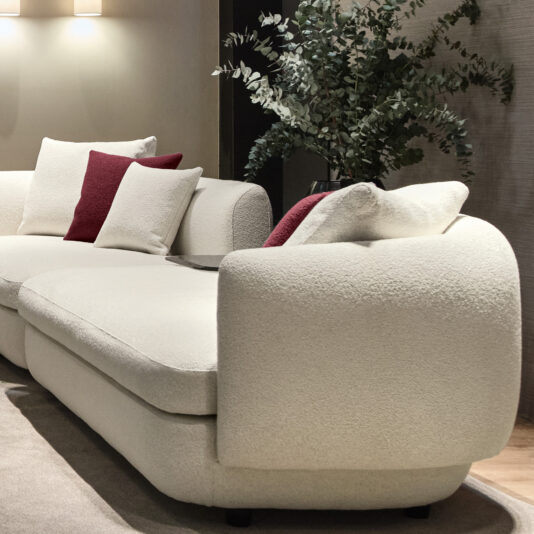 Large Modular Contemporary Corner Sofa A modern living room highlights a large Modular Contemporary Corner Sofa in cream, decorated with plush white and dark red cushions. Beside it stands a lush green plant, complemented by gentle overhead lighting that sets a cozy ambiance. A neutral-toned rug rests beneath the arrangement.