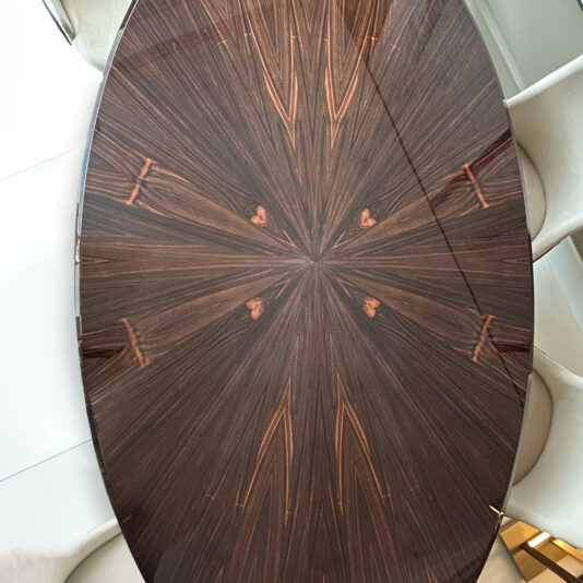 The Exclusive Oval Designer Dining Set features a glossy dark wood table with a starburst pattern, surrounded by beige upholstered chairs, offering a modern, elegant, and sophisticated dining experience.