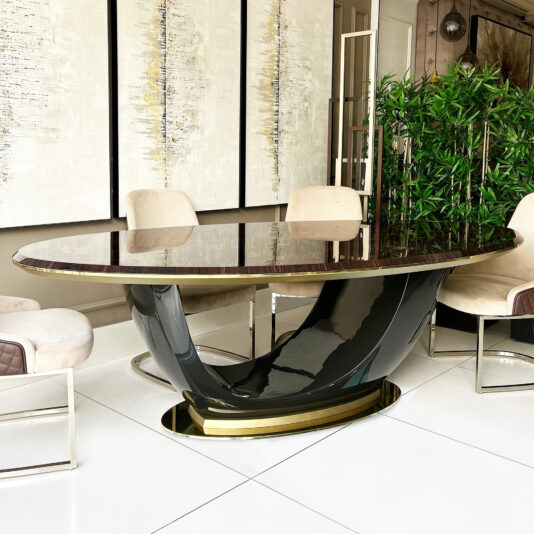A modern dining room showcases the Exclusive Oval Designer Dining Table with a sculptural black and gold base, cream upholstered chairs, abstract wall art behind it, and a tall indoor plant for added elegance.