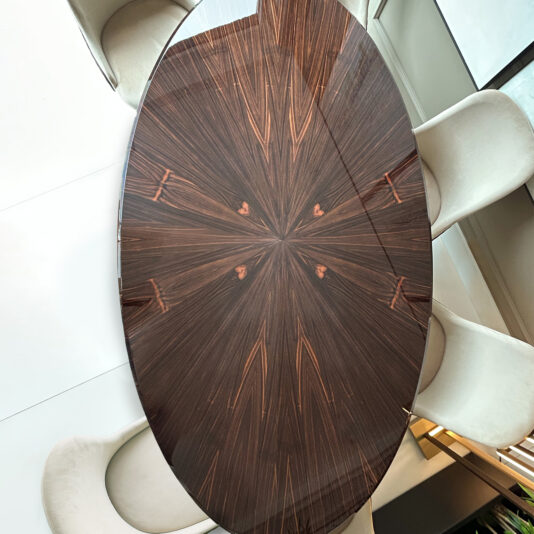The Exclusive Oval Designer Dining Table boasts a glossy dark wood surface with a starburst pattern, paired with six light-coloured cushioned chairs in a modern, well-lit space.