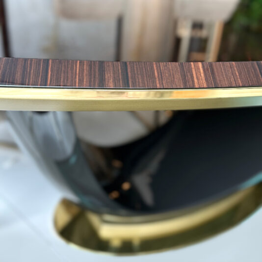 A close-up of the Exclusive Oval Designer Dining Table shows its wooden top with horizontal dark and light stripes, gold-coloured trim, and a curved glossy black support base.
