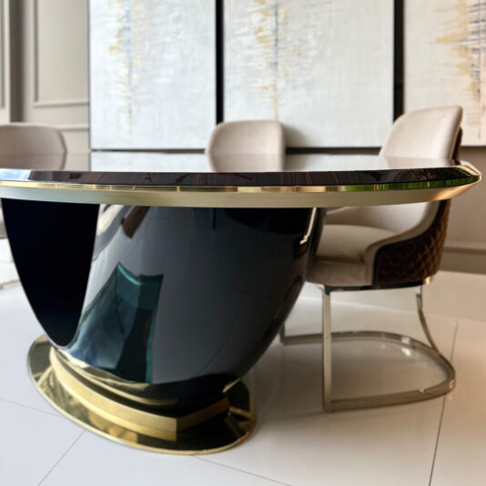 The Exclusive Oval Designer Dining Table with a glossy black and gold base pairs with beige cushioned chairs, whilst abstract art on the wall completes the sleek, contemporary setting.