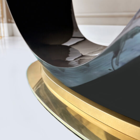 A close-up of the Exclusive Oval Designer Dining Table, featuring a glossy black curved form on a gold and glass base, is set against a minimalistic white background.
