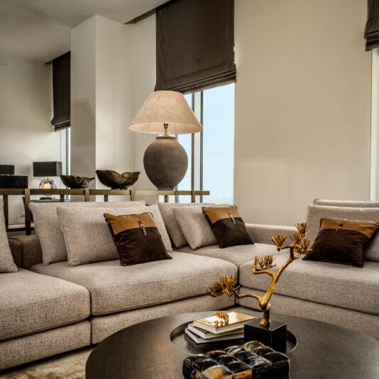 Modern living room with a large grey sectional sofa, brown accent cushions, a round dark wood coffee table decorated with books and a gold branch ornament, and a contemporary table lamp near tall windows with brown blinds.
