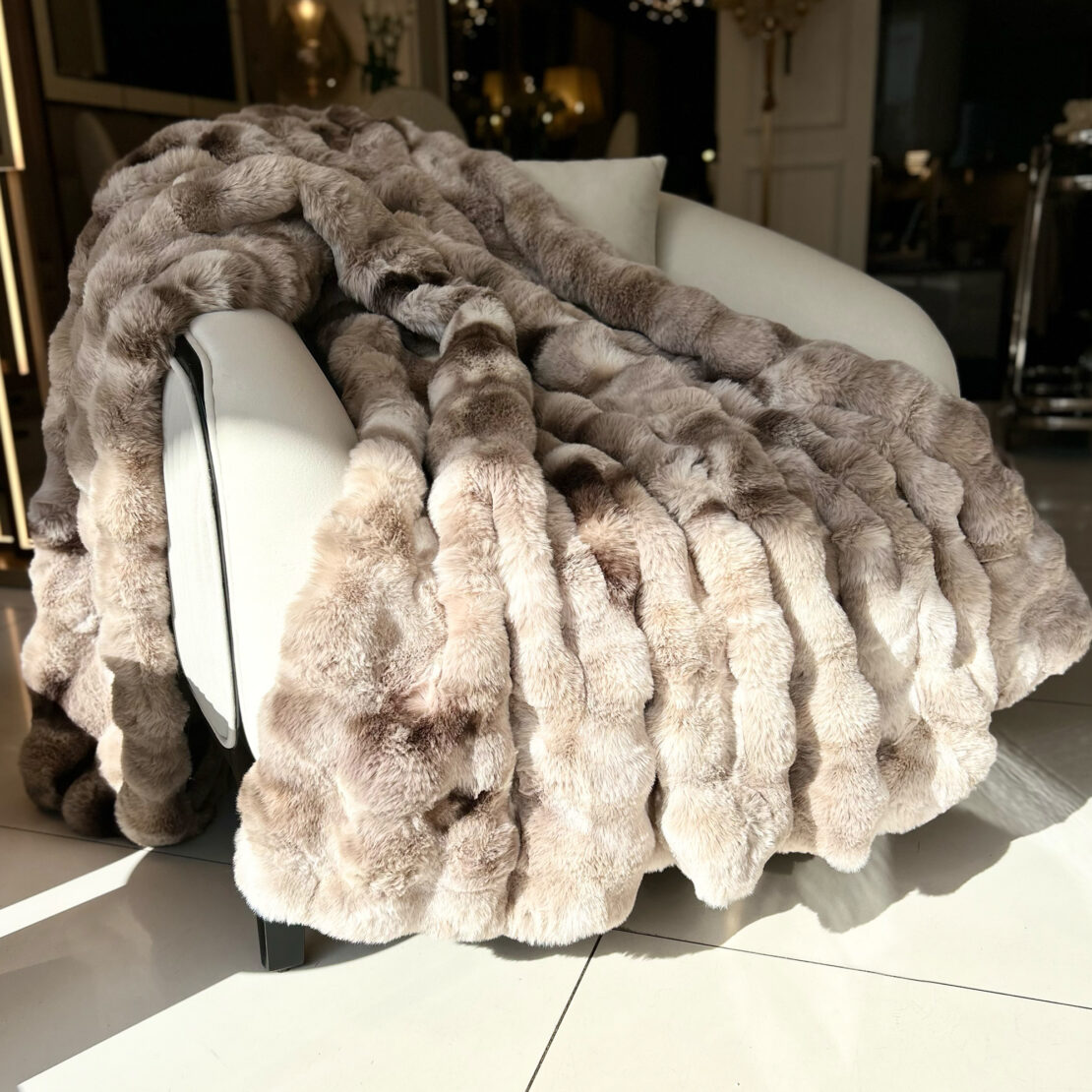 A Beige Textured Faux Fur Throw in light brown hues adorns a white armchair, with sunlight casting soft shadows on the smooth tiled floor, enhancing the room's cozy and elegant ambiance.