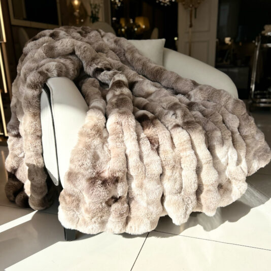 A Beige Textured Faux Fur Throw in light brown hues adorns a white armchair, with sunlight casting soft shadows on the smooth tiled floor, enhancing the room's cozy and elegant ambiance.