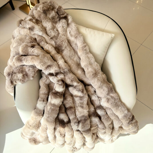 A Beige Textured Faux Fur Throw drapes over a white modern armchair with a cream cushion, set on a tiled floor basking in natural light.