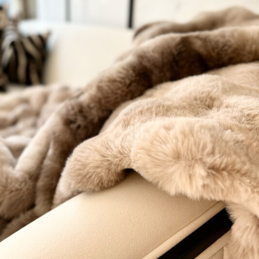 A close-up shows the Beige Textured Faux Fur Throw, soft and plush, draped over a light-colored couch, appearing fluffy and luxurious with a zebra-print cushion in the blurred background.