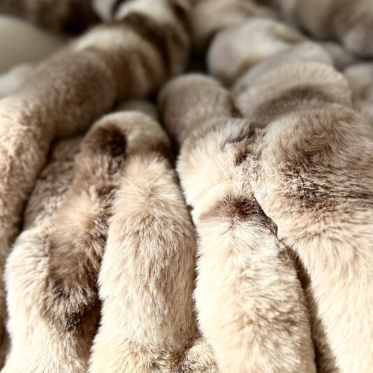 A close-up of the Beige Textured Faux Fur Throw reveals its luxurious plush texture in brown shades. The fluffy, inviting appearance and visible fabric folds enhance its opulent look, perfect for cozy elegance.