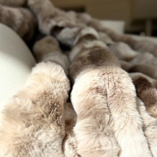 A Beige Textured Faux Fur Throw is elegantly draped over a light surface, with sunlight casting gentle shadows that highlight its plush texture and warm hues.