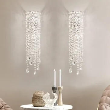 Contemporary Cut Crystal Wall Light The beige wall is adorned with a Contemporary Cut Crystal Wall Light that casts a shimmering glow. Below it, the table displays a woven vase, a sculpted glass bowl, and two candlesticks with unlit white candles—one gold and one silver.