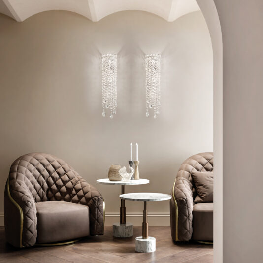 A cozy seating area features two plush, quilted brown armchairs on a wooden floor with marble side tables in between, holding candles and decor. Contemporary Cut Crystal Wall Lights elegantly adorn the soft beige wall.