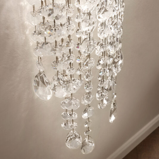 A close-up of the Contemporary Cut Crystal Wall Light showcases faceted glass pieces reflecting light. The varying shapes of the crystals, attached to metal fixtures, set against a light wall background, create a stunning interplay of light and elegance.