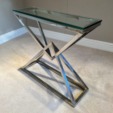 Contemporary Stainless Steel Glass Console Table The Contemporary Stainless Steel Glass Console Table features a zigzag chrome frame with a gold finish, adding modern flair. Its clear glass top and angular design stand elegantly on a carpeted floor against a plain wall.