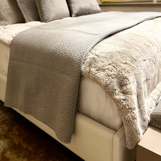 A neatly made bed showcases the Large Silver Quilted Throw elegantly draped over a fluffy cream fur. The ensemble includes multiple matching pillows, with the neutral-colored bed frame complementing the warm-toned floor perfectly.