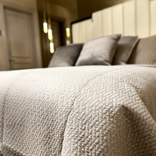 A neatly made bed features a textured light gray quilt, enhanced by the elegant Large Silver Quilted Throw. Several pillows in complementary colors rest against the headboard, and pendant lights softly illuminate the room in the background.