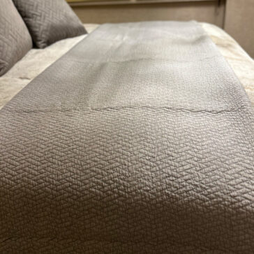 Close-up of a neatly made bed with beige bedding and a gray textured runner, topped with the Large Silver Quilted Throw. Patterned pillows add flair, while soft lighting creates a cozy ambiance.