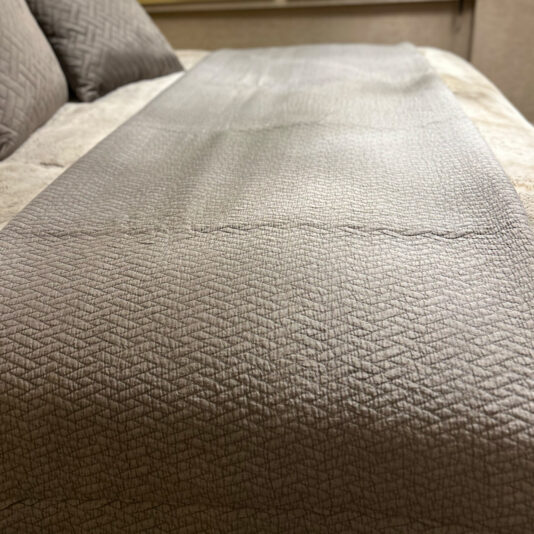 Close-up of a neatly made bed with beige bedding and a gray textured runner, topped with the Large Silver Quilted Throw. Patterned pillows add flair, while soft lighting creates a cozy ambiance.