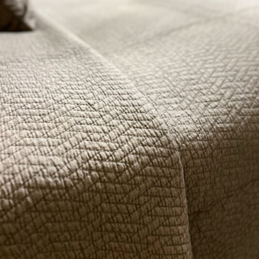 A close-up of a textured beige quilted bedspread features a zigzag pattern, complemented by the Large Silver Quilted Throw. The soft, slightly wrinkled fabric covers the bed, with gentle lighting highlighting the intricate stitching of both layers.