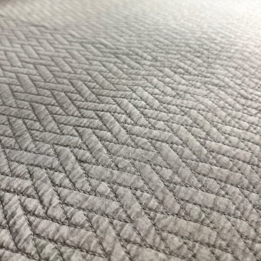 The Large Silver Quilted Throw features light gray textured fabric with a herringbone pattern and uniform stitching, accentuating its subtle sheen under lighting.