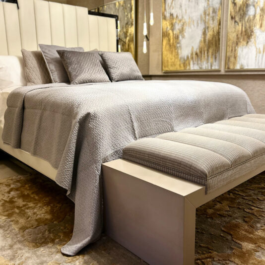 A luxurious bedroom showcases a neatly made bed with a light gray quilt and pillows. At the foot sits a padded bench draped with the Large Silver Quilted Throw. Gold and silver abstract art adorns the wall, enhancing the elegant neutral color scheme.