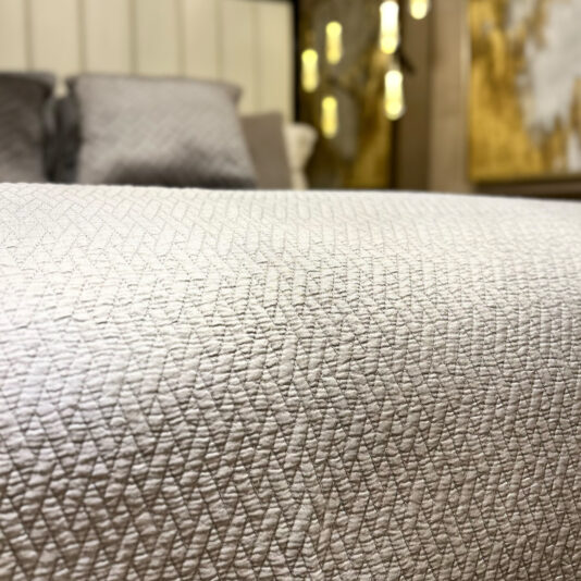 A close-up shows a textured beige bedspread on a bed featuring the Large Silver Quilted Throw. The headboard and plush pillows amplify the warmth, while soft lighting and cozy hues make for an inviting ambiance.