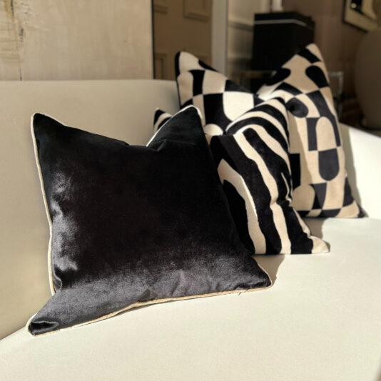 A close-up of a white couch features luxury velvet scatter cushions, including a plush black double-sided design at the front. Behind it are two cushions with bold black and white geometric patterns, showcasing modern elegance.