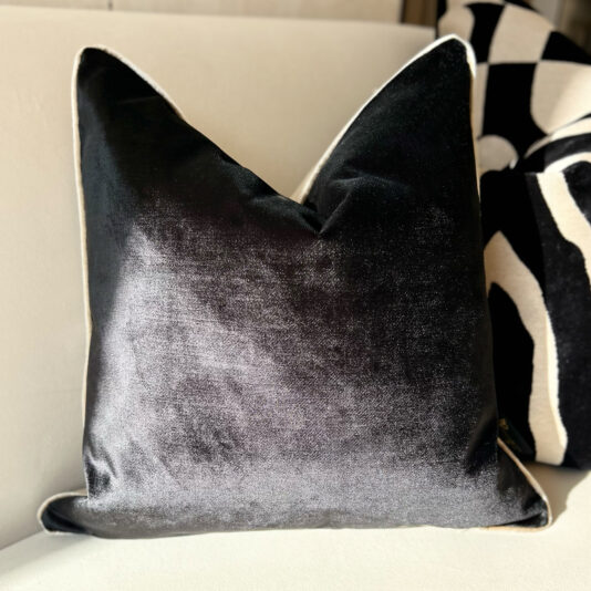 A Luxury Velvet Double Sided Scatter Cushion with a subtle sheen is on a light-colored couch, with a patterned black and white pillow behind it for contrast.