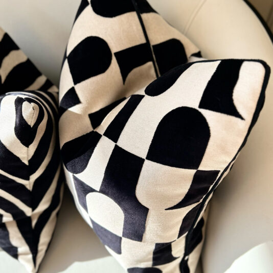Two Luxury Velvet Retro Pattern Scatter Cushions, showcasing a black and white geometric pattern with large shapes and curves, rest elegantly on a light-colored seat in soft natural light.