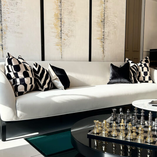 A modern living room has a white sofa with Luxury Velvet Retro Pattern Scatter Cushions in black and white. A circular black coffee table displays a silver and gold chess set, while abstract wall art with retro vertical lines serves as the backdrop.