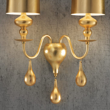 A golden wall sconce with two lampshades is mounted on a textured gray wall, featuring elegant curved arms and drop-shaped accents. Reminiscent of the Modern Gold Leaf Pendant Light (Copy), it exudes a luxurious and ornate appearance.