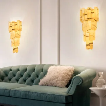 Modern Onyx Tiered Wall Light A plush green velvet sofa with a fluffy beige pillow sits in front of a white wall. Two elegant amber sconces cast soft light, complemented by the Modern Onyx Tiered Ceiling Light above. A small white side table holds a clear glass carafe and glass.