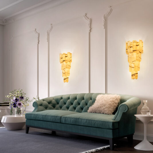 A luxurious living room showcases a tufted teal sofa with a fluffy gray pillow. Two golden wall sconces and the Modern Onyx Tiered Ceiling Light illuminate the white paneled walls warmly. A round coffee table holds a vase of flowers and a small container, enhancing the elegance.