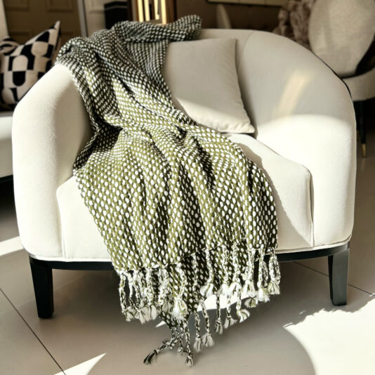 A white chair with wooden legs is adorned with the Olive Green Throw With Tassels, accompanied by two white pillows, all placed on a light-colored tiled floor in a cozy room.