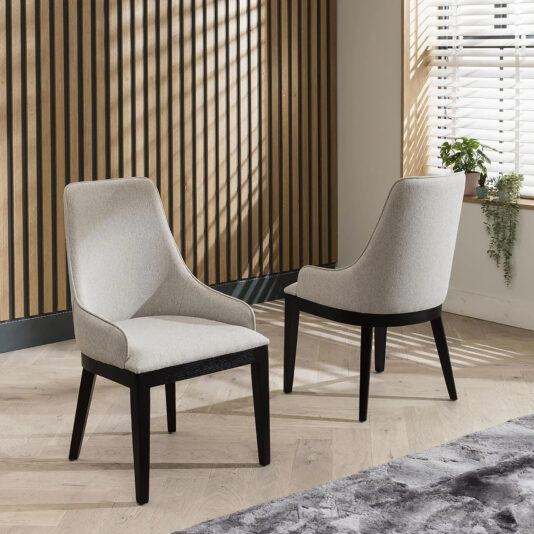 On a light wood floor, a Set Of 6 Beige High Back Dining Chairs is arranged, with two featuring dark wooden legs—one facing forward, the other backward. Vertical wooden slats and blinds cast intriguing shadows in the background alongside a small plant.