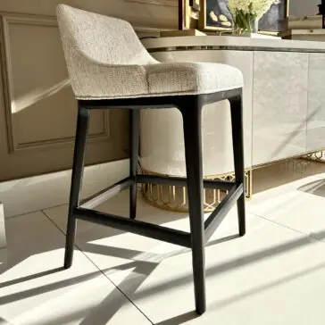 A sleek Set Of Two Boucle Bar Stools with light fabric seats and dark wooden legs is stylishly set on a glossy tiled floor. Sunlight casts their shadows, accentuated by a chic white cabinet and elegant decor in the background.