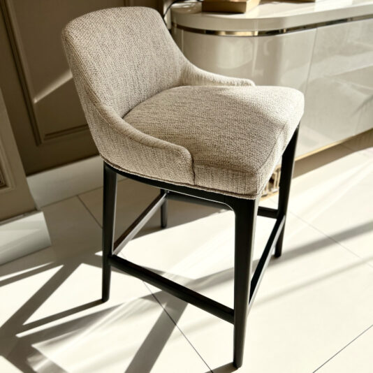 Set Of Two Boucle Bar Stools Part of the Set of Two Boucle Bar Stools, a beige upholstered bar stool with a curved backrest and black wooden legs sits on a glossy tiled floor. Sunlight highlights its texture, while a sleek cabinet is partially visible in the background.