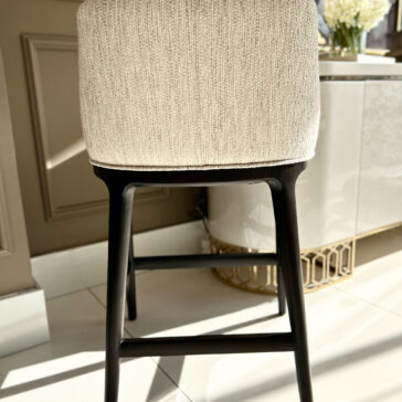 A modern Set Of Two Boucle Bar Stools, featuring beige boucle fabric seats and black legs, stands on a white tiled floor. In the background, a white cabinet with a gold decorative base is partially visible as sunlight creates inviting shadows across the room.