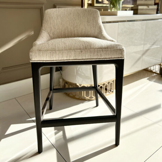 Set Of Two Boucle Bar Stools A beige upholstered chair with a high backrest and black wooden legs stands on a tiled floor. Sunlight casts shadows from the chair and surrounding items, including the Set Of Two Boucle Bar Stools nearby. A cabinet is partially visible in the background.