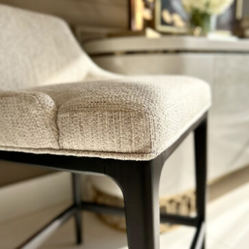 Close-up of a modern chair with a textured beige fabric cushion and dark wooden legs. A blurred background reveals a white sideboard, framed picture, and the Set Of Two Boucle Bar Stools, suggesting a stylish interior setting.